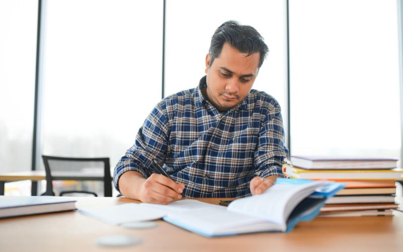 A man sitting at a desk, engrossed in a book, with a pen in hand, deep in thought and concentration.